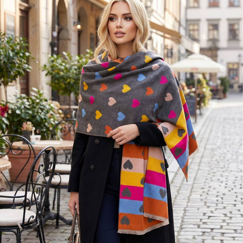 A woman modeling a dark gray knit wrap with colorful heart motifs and striped sections featuring hearts, photographed on a cobblestone street.