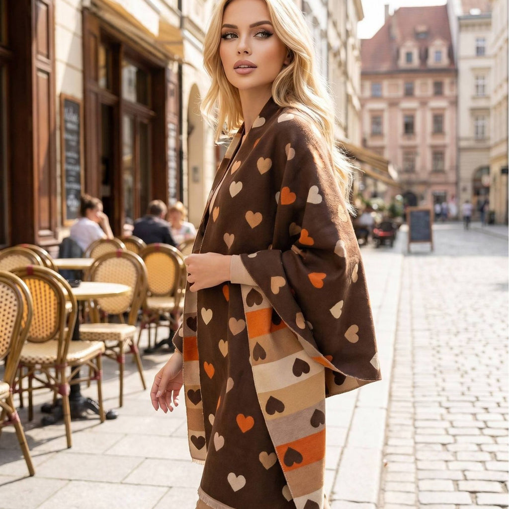 A woman stands on a cobblestone street in front of a cafe, wearing a brown kimono-style cardigan with an all-over heart pattern in beige, orange, and dark brown.