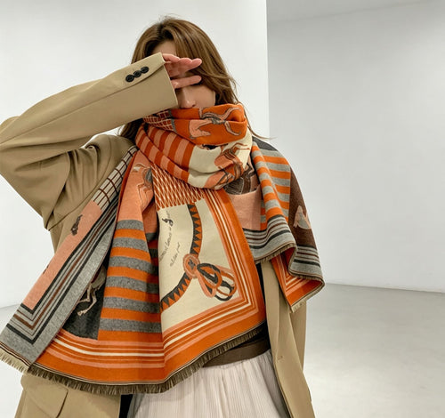 A woman wearing a beige blazer and white skirt models a large orange, beige, and gray patterned scarf with fringe.