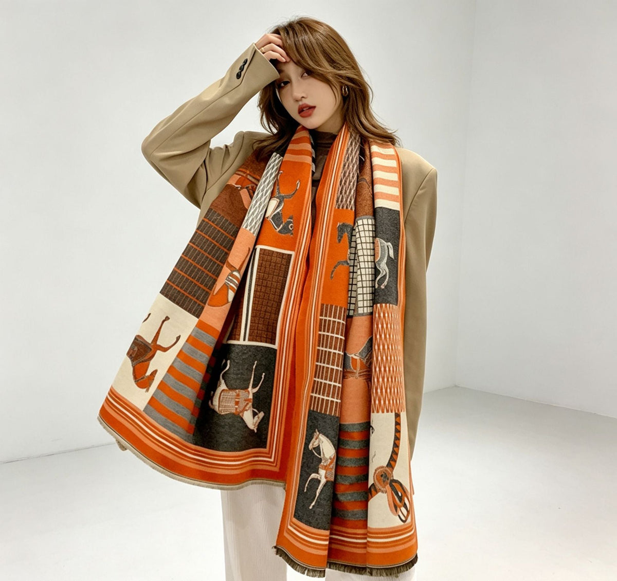 A woman models a long, wide scarf with a patchwork design featuring orange, brown, beige, and black blocks with animal and geometric motifs, styled over a beige blazer and white pants.