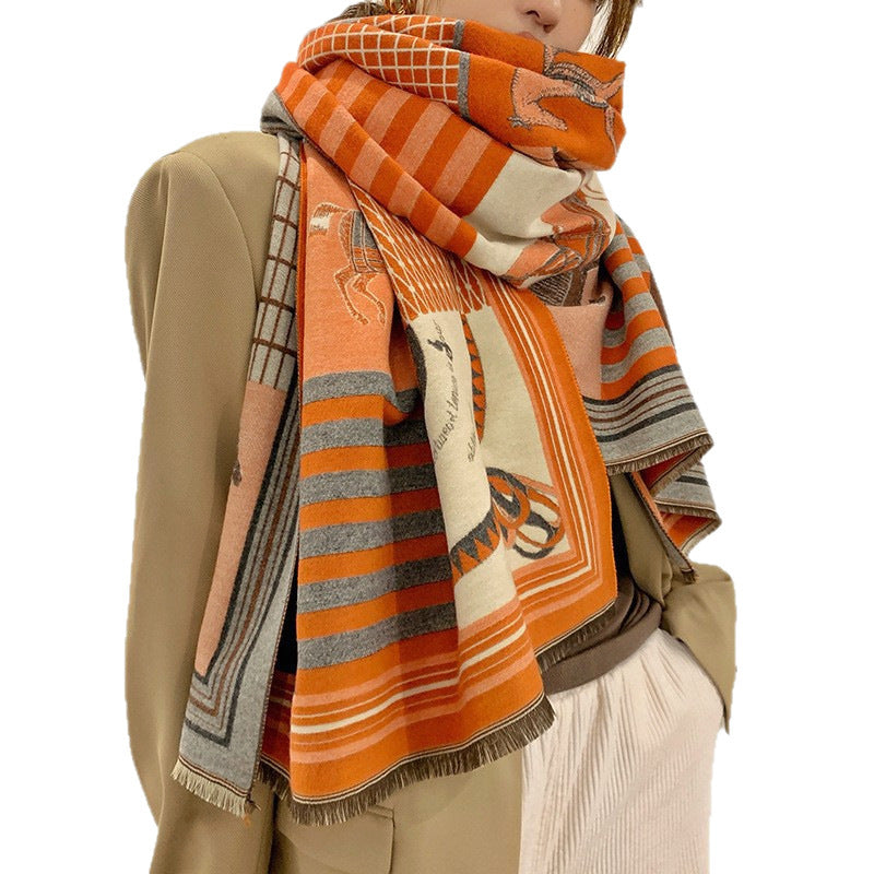 A person wearing a large orange, beige, and gray patterned scarf wrapped around their neck, styled with a beige blazer and white pants against a plain white background.