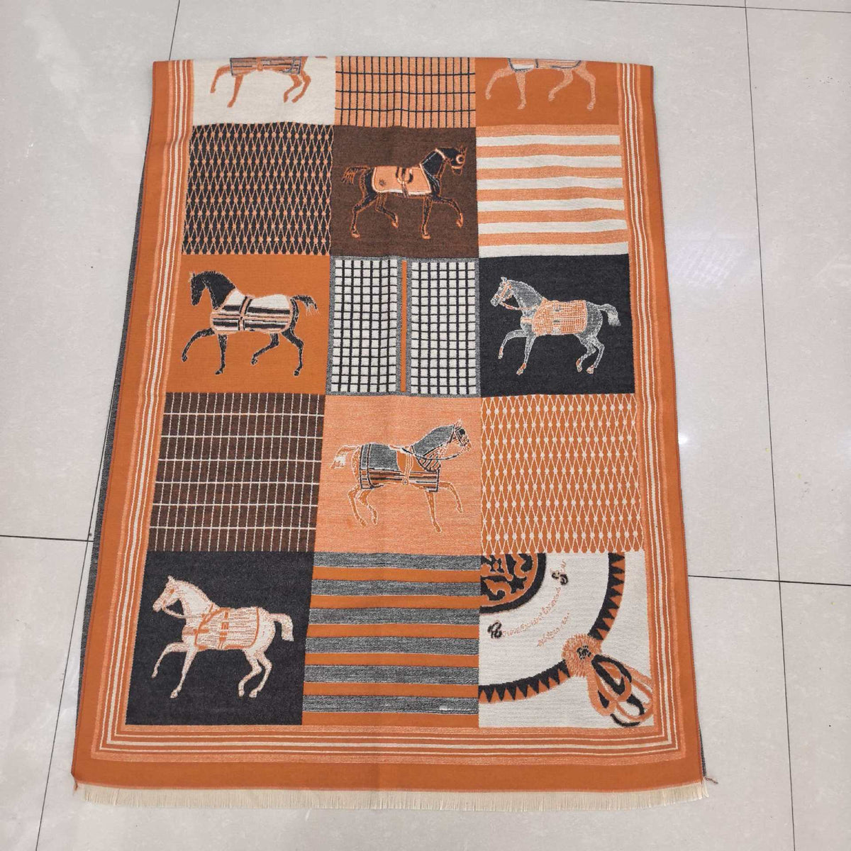 A rectangular woven throw blanket with a patchwork design featuring horses and geometric patterns in orange, brown, black, and cream, laid flat on a light tiled floor.