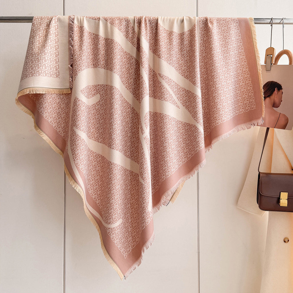 A large square scarf in dusty pink and cream hangs from a metal rack on a wall. The scarf features a fine geometric pattern, fringed edges, and a wide cream border.
