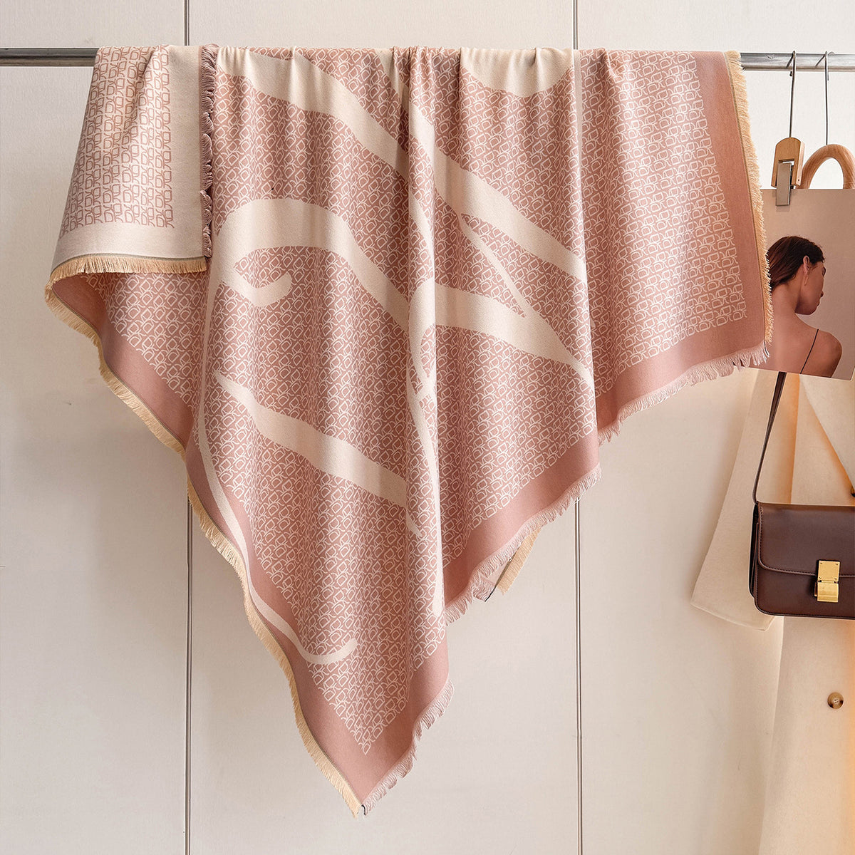 A large square scarf in dusty pink and cream hangs from a metal rack on a wall. The scarf features a fine geometric pattern, fringed edges, and a wide cream border.