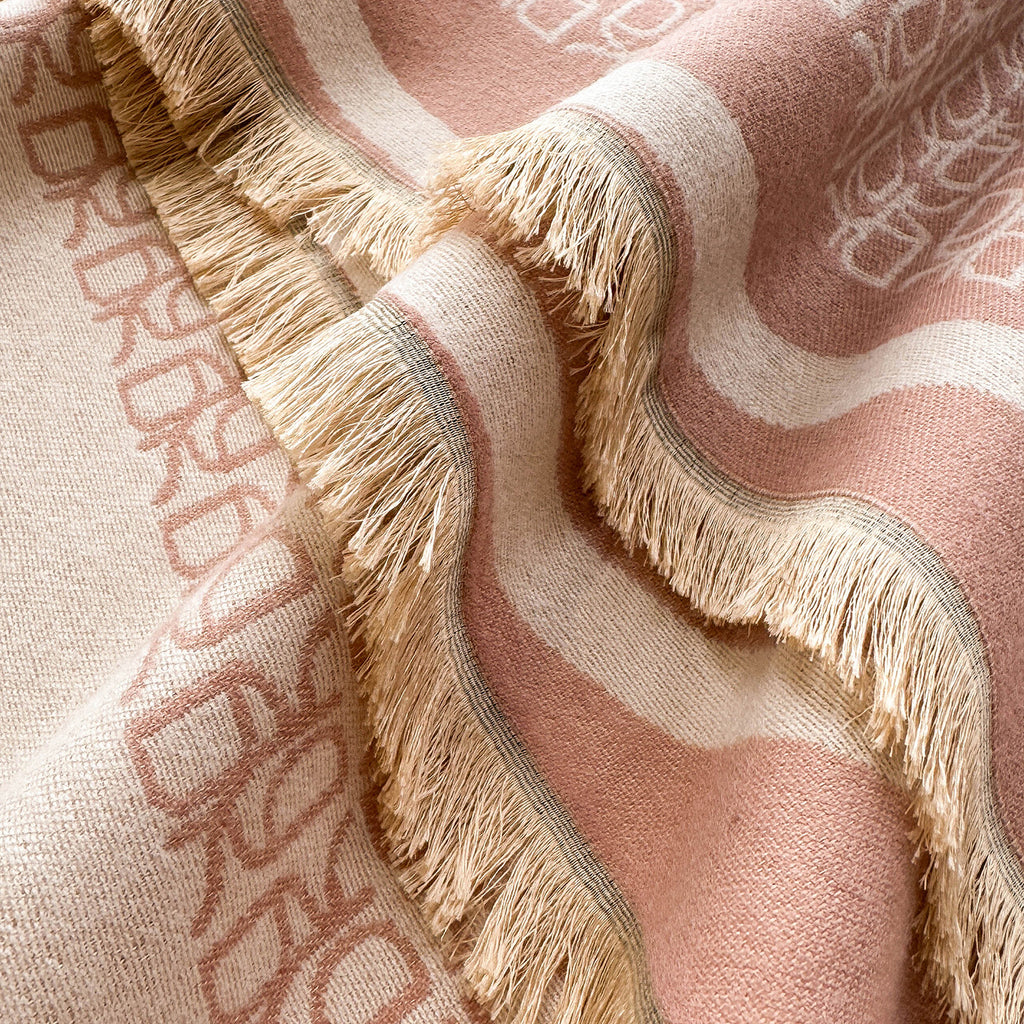 A close-up of a woven throw blanket with fringe, featuring a dusty pink and cream striped pattern with the brand name 'BURBERRY' woven into the fabric.