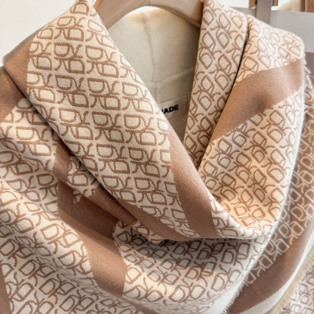 A close-up of a beige and cream scarf draped on a hanger, featuring a repeating geometric monogram pattern.