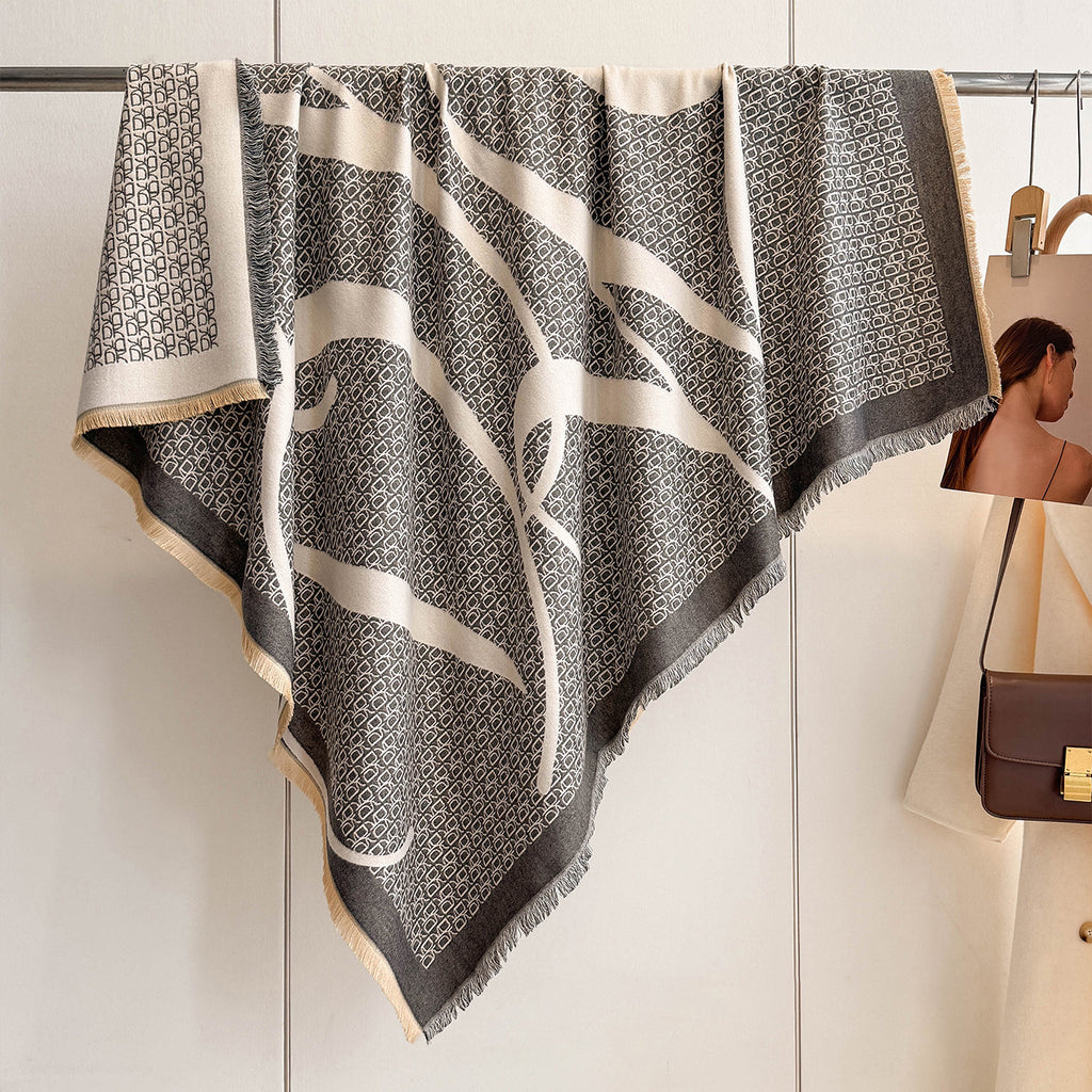 A large square scarf with a black and beige geometric pattern and fringe, hanging from a metal rack against a white wall. A small fashion magazine and a brown handbag are partially visible on the right side.