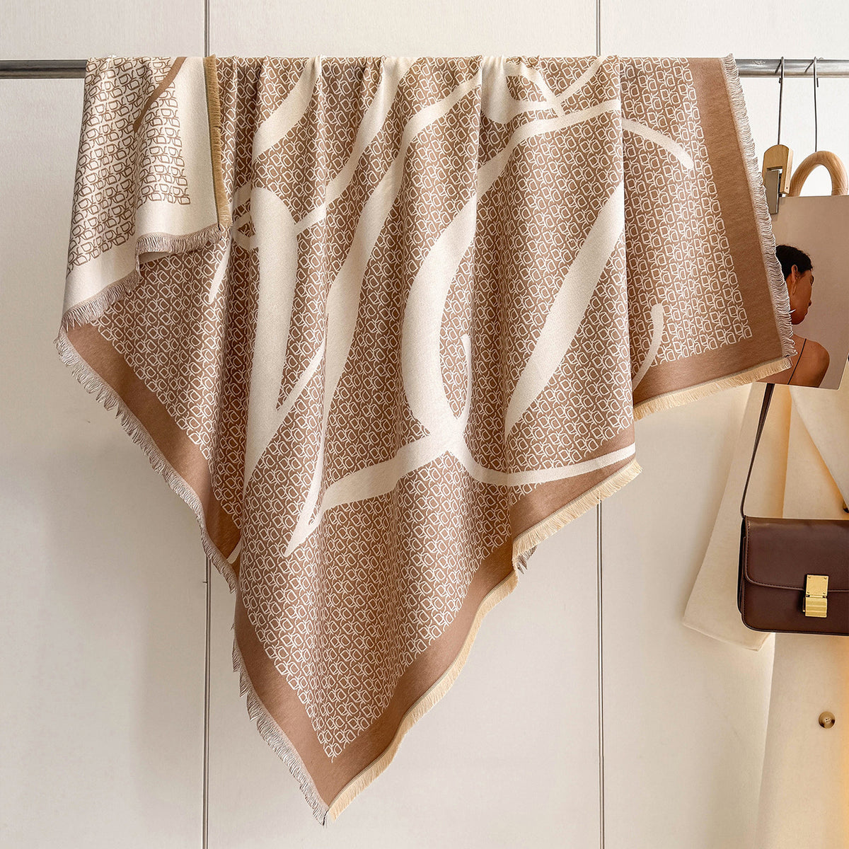 A beige and white patterned scarf with fringe, draped over a clothing rack in a styled interior setting.