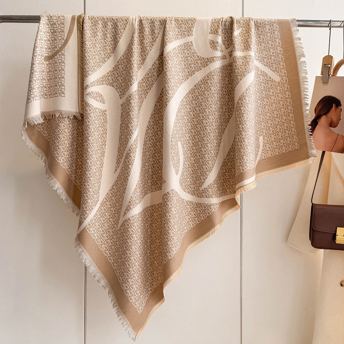 A large beige and cream patterned scarf with fringe edges hangs from a metal rack against a light wall. A partial model in a cream coat and brown shoulder bag is visible on the right side.
