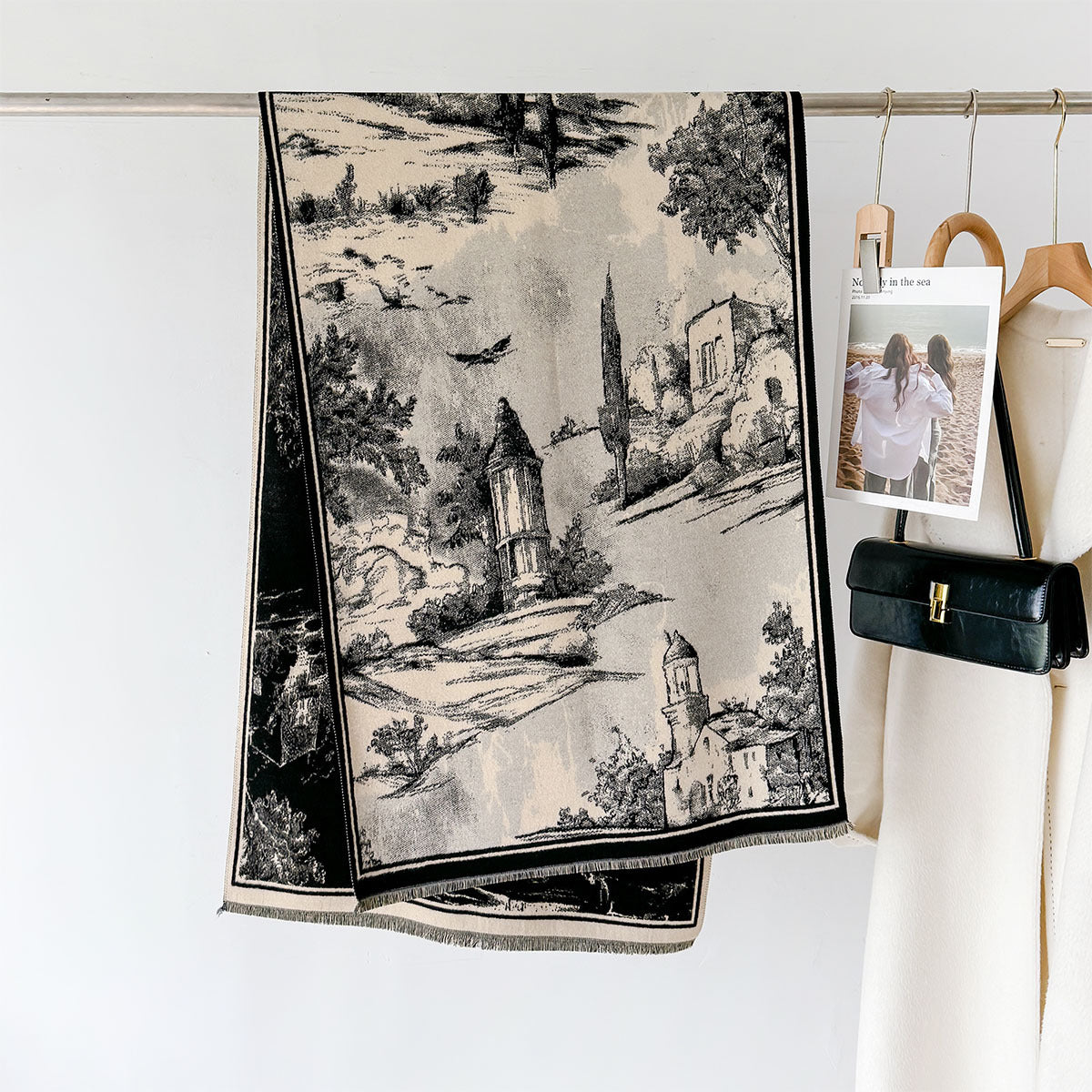 A black and beige woven scarf with a scenic landscape print hangs from a metal rod on a white wall, styled next to a cream-colored coat and a black handbag.