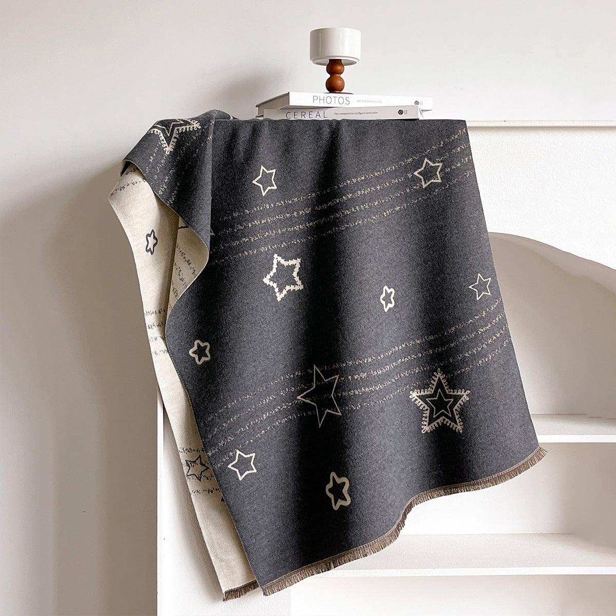 A dark charcoal gray throw blanket with a pattern of cream stars and horizontal streaks, draped over a white shelf next to a small white lamp and books.
