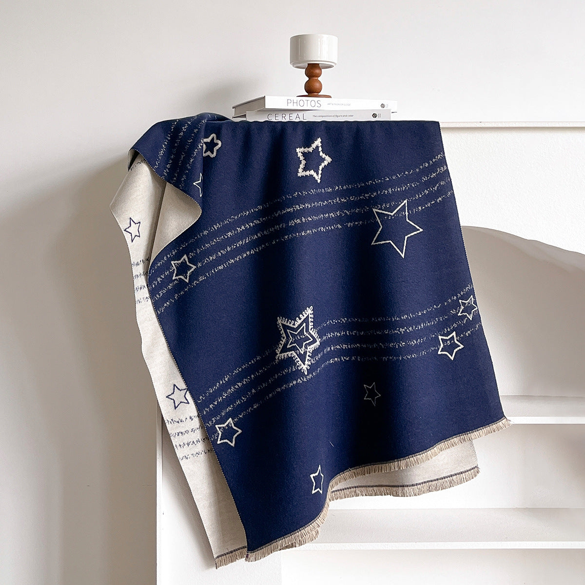 A navy blue throw blanket with white embroidered stars and stripes, draped over a white wooden shelf with a small wooden lamp on top.