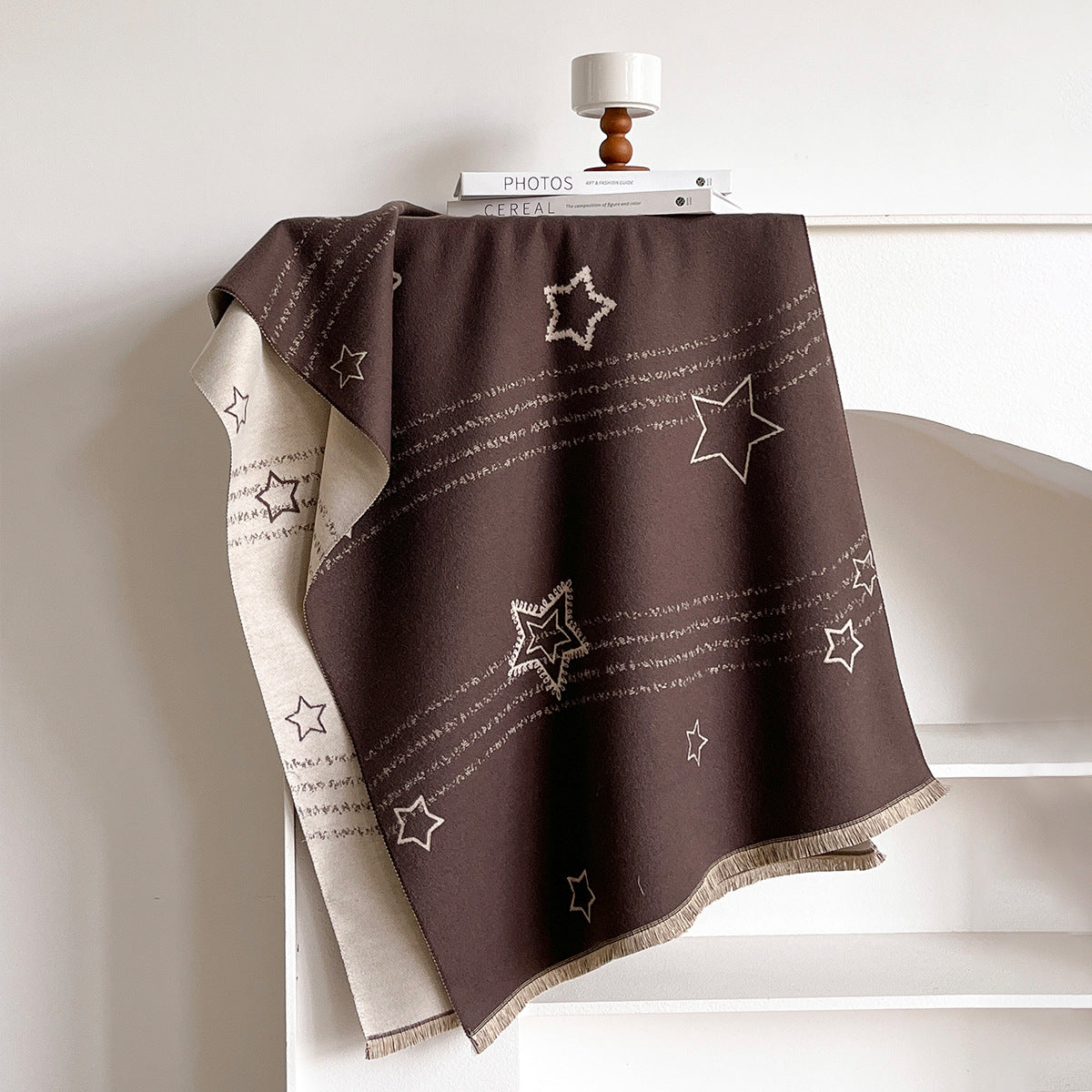 A dark brown throw blanket with white embroidered stars and stripes draped over a white shelf, showing a cream reverse side with text patterns.