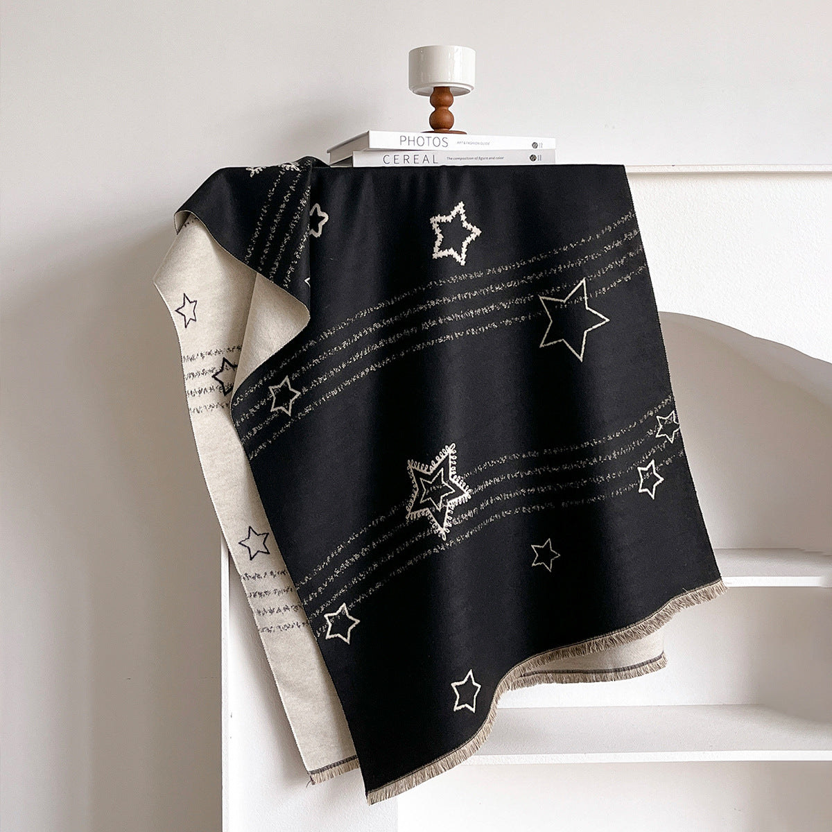 A black throw blanket with white star and stripe patterns draped over a white shelf, with a cream reverse side and fringe visible.