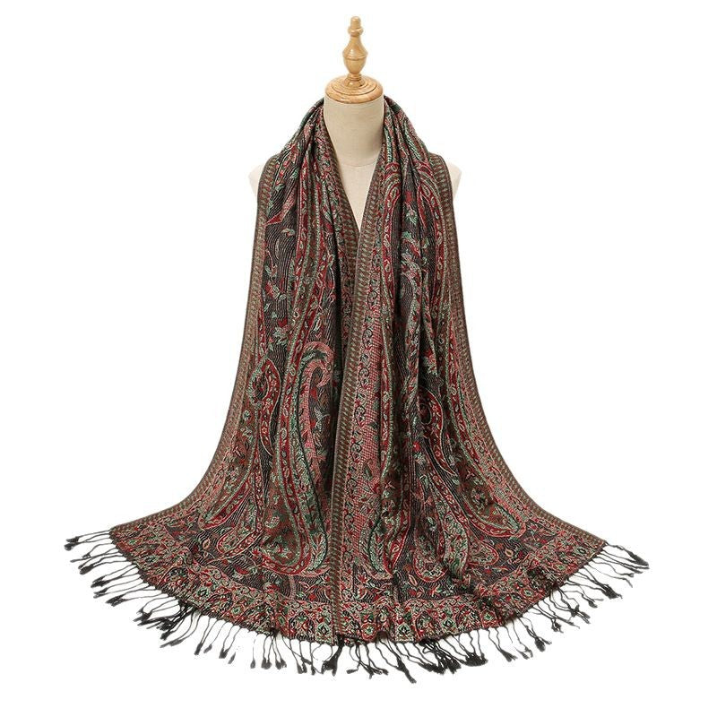 A long paisley scarf draped on a mannequin torso against a plain white background. The scarf has a dark brown base with intricate red, green, and beige paisley patterns and black fringe tassels along the edges.