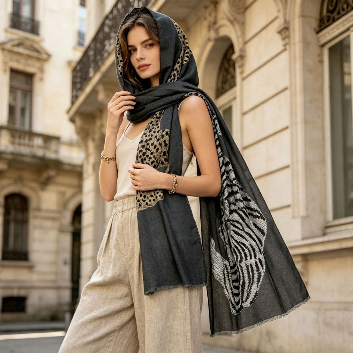 A woman wearing a black and beige animal-print hooded scarf with a tiger-patterned lower panel, styled over a white camisole and beige linen pants in front of a classic stone building.