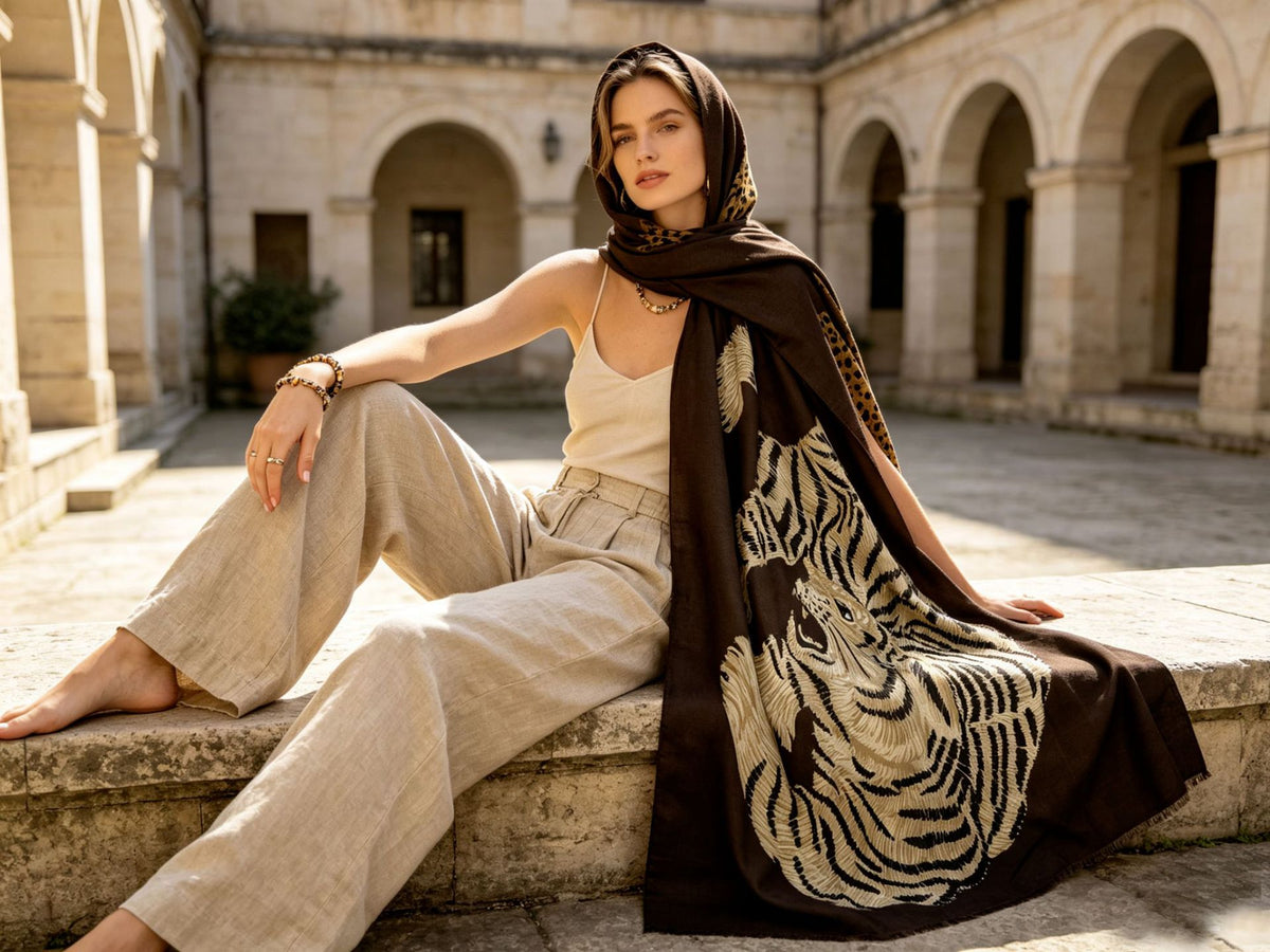 A woman sits on a stone ledge in a sunlit courtyard, wearing a beige camisole, wide-leg beige trousers, and a dark brown headscarf with a large tiger-stripe pattern and gold-toned chain details.